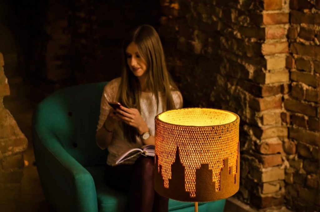 dziewczyna siedząca na fotelu dzwoni do przyjaciółki, a na stoliku stoi designerska lampa z motywem miasta